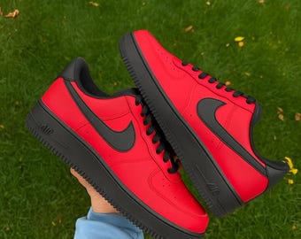May include: Pair of red sneakers with black accents, including the Nike swoosh logo, laces, and sole. The shoes are displayed against a green grassy background. The sneakers are a classic athletic style.