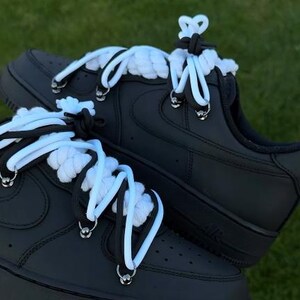 K&ouml;nnte beinhalten: Schwarze AF1 "Black Panda" Sneaker mit wei&szlig;en und schwarzen Seilschn&uuml;rsenkeln. Die Schuhe haben eine schwarze Sohle und ein schwarzes Obermaterial mit silbernen &Ouml;sen. Der Text "AF1 "BLACK PANDA" steht oben im Bild.