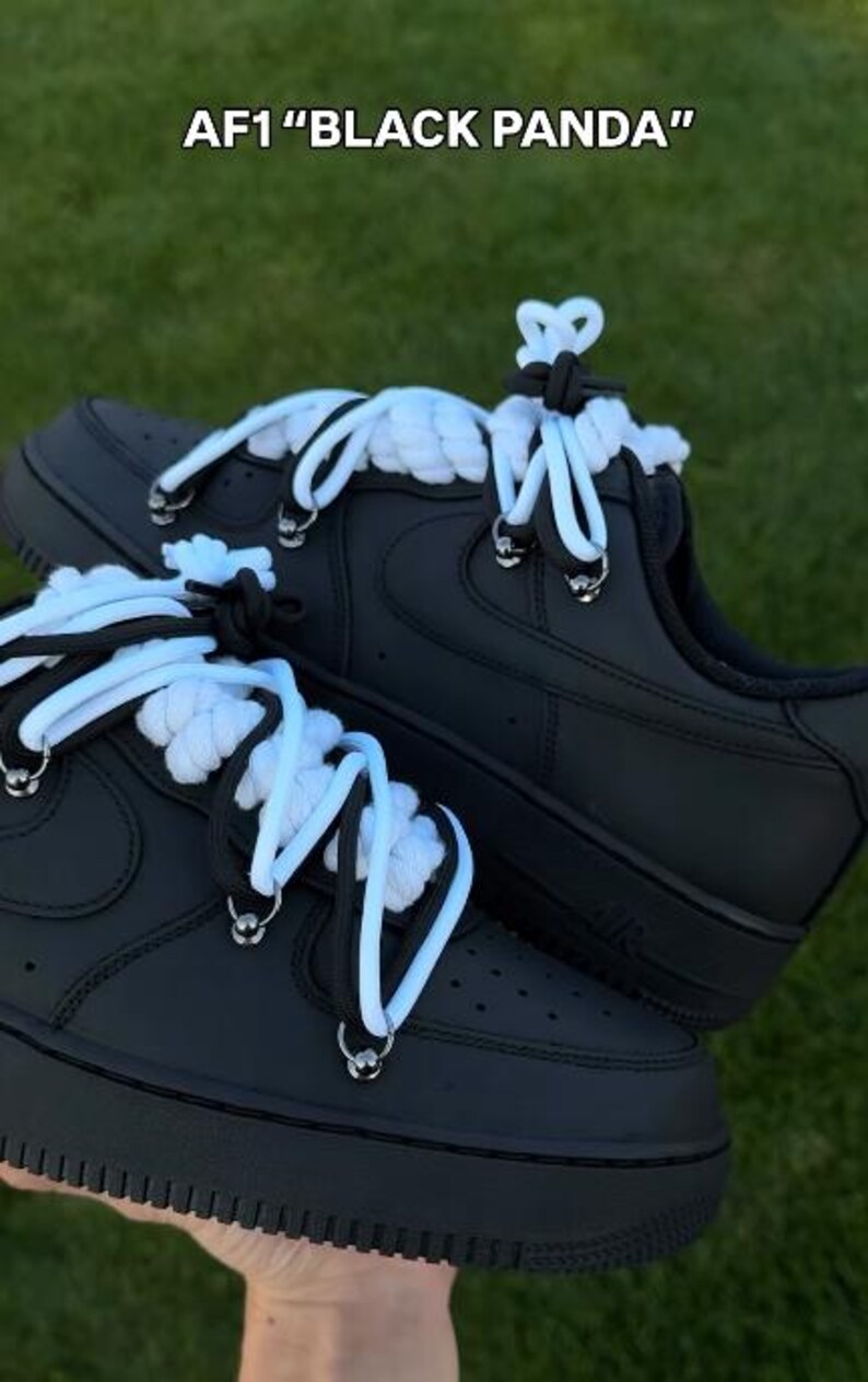 K&ouml;nnte beinhalten: Schwarze AF1 "Black Panda" Sneaker mit wei&szlig;en und schwarzen Seilschn&uuml;rsenkeln. Die Schuhe haben eine schwarze Sohle und ein schwarzes Obermaterial mit silbernen &Ouml;sen. Der Text "AF1 "BLACK PANDA" steht oben im Bild.