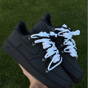K&ouml;nnte beinhalten: Paar schwarze AF1 "Black Panda" Sneaker mit wei&szlig;en Schn&uuml;rsenkeln. Die Schuhe werden vor einem gr&uuml;nen Hintergrund gehalten. Der Text "AF1 "BLACK PANDA"" befindet sich oben im Bild.