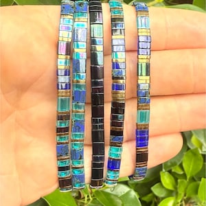 May include: Five beaded bracelets in shades of blue, teal, and black with gold-colored accents. The bracelets are made of small, rectangular beads and are displayed against a background of green foliage.