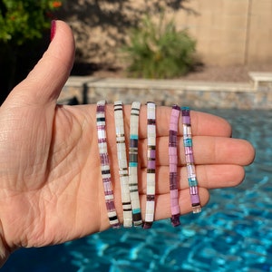 May include: Six colorful beaded bracelets with a mix of white, purple, and turquoise beads. The bracelets are stacked on top of each other and are held in a hand.