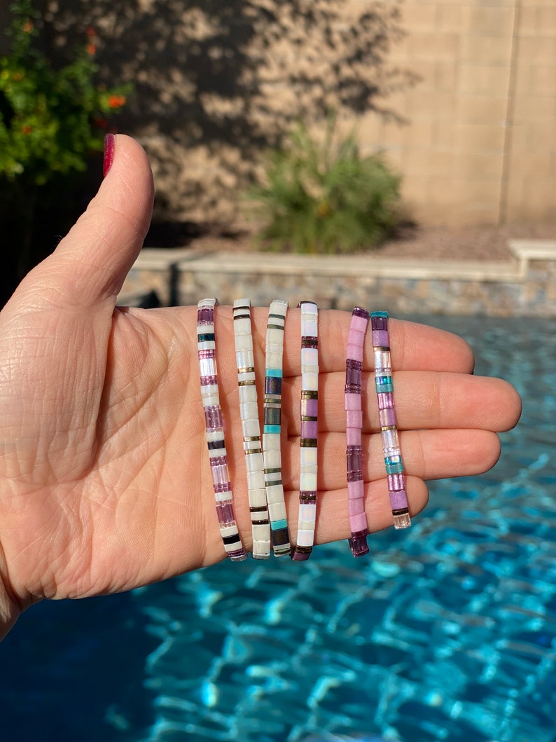 May include: Six colorful beaded bracelets with a mix of white, purple, and turquoise beads. The bracelets are stacked on top of each other and are held in a hand.
