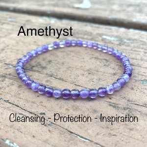 May include: Amethyst bracelet made with small purple beads. The bracelet is on a wooden surface. The text "Cleansing - Protection - Inspiration" is below the bracelet.