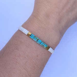May include: A white bracelet with a turquoise and gold accent. The bracelet is made of small square tiles.