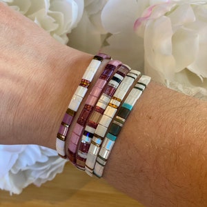 May include: A stack of five colorful tile bracelets on a person's wrist. The bracelets are made of white, pink, blue, and gold tiles.