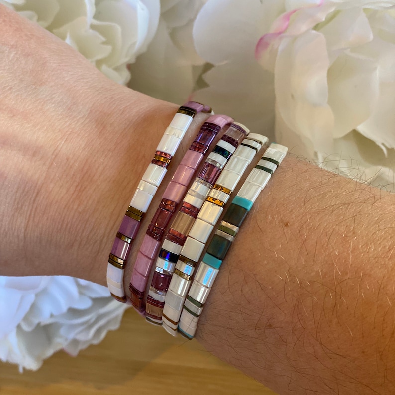 May include: A stack of five colorful tile bracelets on a person's wrist. The bracelets are made of white, pink, blue, and gold tiles.