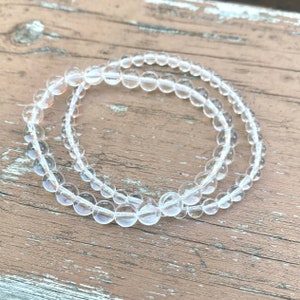 May include: Two clear crystal beaded bracelets on a wooden surface. The bracelets are made of small, round beads and are stacked on top of each other.