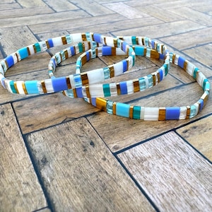 May include: Three stacked bracelets with a white, blue, and gold color scheme. The bracelets are made of small square tiles.