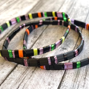 May include: Three colorful bracelets made of black, green, orange, and purple square beads. The bracelets are stacked on top of each other.