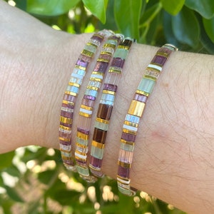 May include: Four stacked bracelets with a mix of purple, white, and gold beads. The bracelets are made of small, square beads.