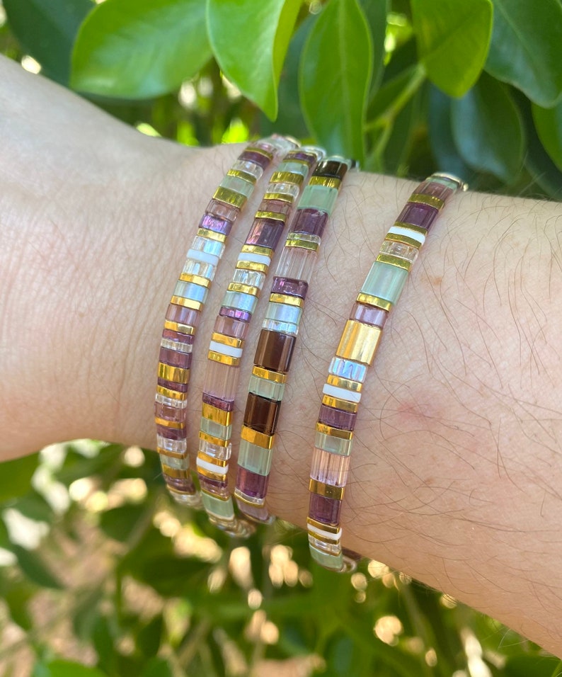 May include: Four stacked bracelets with a mix of purple, white, and gold beads. The bracelets are made of small, square beads.