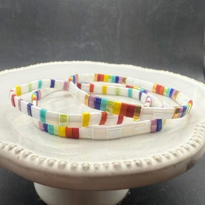 May include: Three white tile bracelets with colorful rainbow accents. The bracelets are stacked on a white ceramic plate. The bracelets have a rectangular tile design with a variety of colors.