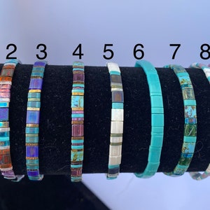 May include: Nine turquoise and multi-colored tile bracelets displayed on a black stand. The bracelets are numbered B-1 through 9.