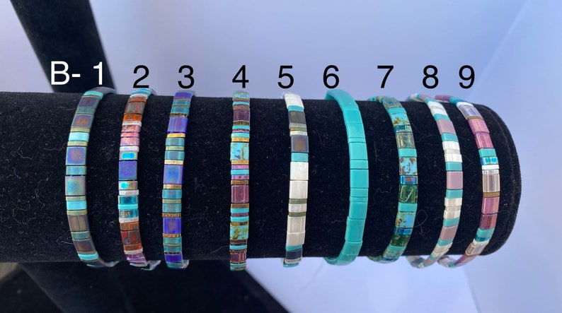 May include: Nine turquoise and multi-colored tile bracelets displayed on a black stand. The bracelets are numbered B-1 through 9.
