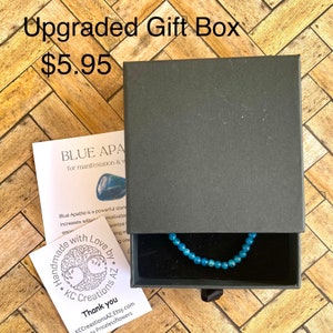 May include: A black gift box with a blue apatite bracelet inside. The box has a label that reads "BLUE APATITE for manifestation & ... Handmade with Love by KC Creations AZ." The label also includes a tree with roots and branches. The text "Thank you" is printed below the tree. The text "KCCreationsAZ.Etsy.com by Pricelessflowers" is printed below the "Thank you" text.