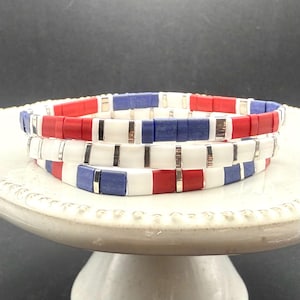 May include: Three stacked bracelets with a patriotic color scheme. Each bracelet features a repeating pattern of red, white, and blue rectangular beads, separated by silver-colored metal bars. The bracelets are displayed on a white, decorative pedestal.