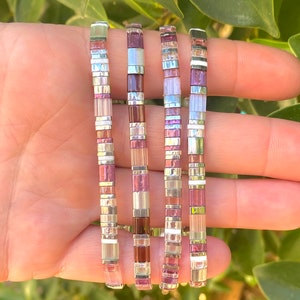 May include: Five colorful bracelets with a mix of pink, white, and silver beads. The bracelets are stacked on top of each other and are made of small, square beads.