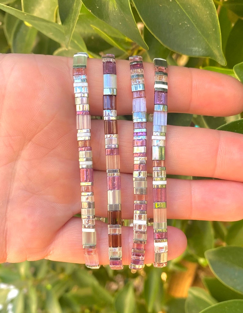 May include: Five colorful bracelets with a mix of pink, white, and silver beads. The bracelets are stacked on top of each other and are made of small, square beads.