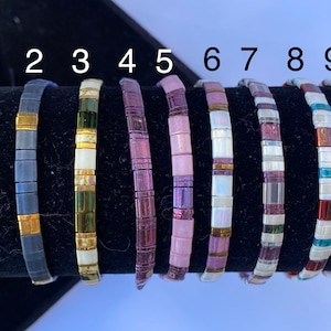 May include: Ten colorful bracelets made of small square tiles. The bracelets are arranged in a row on a black display stand. The bracelets are numbered C-1 through 10.