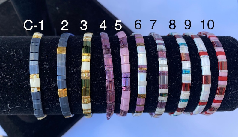 May include: Ten colorful bracelets made of small square tiles. The bracelets are arranged in a row on a black display stand. The bracelets are numbered C-1 through 10.