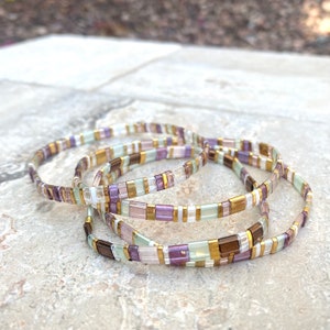 May include: Four stacked bracelets made of small, colorful beads. The bracelets are made of gold, white, purple, and green beads.