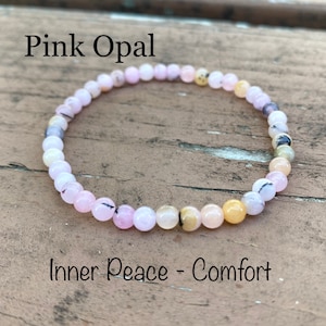 May include: Pink Opal beaded bracelet with a light pink, white, and yellow color scheme. The bracelet is on a wooden surface and the text "Inner Peace - Comfort" is below the bracelet.