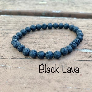 May include: A black lava rock beaded bracelet. The bracelet is made of small, round beads that are all the same size. The beads are a dark gray color and have a rough, textured surface. The bracelet is lying on a wooden surface.