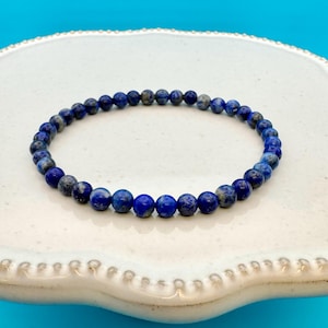 May include: A blue lapis lazuli beaded bracelet with a simple design. The bracelet is made of small, round beads that are strung together on a clear elastic cord.