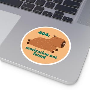 Puede incluir: Una pegatina redonda con un capibara de dibujos animados en tonos marrones y beige, con el texto "404: motivation not found" en verde y naranja. La pegatina está sobre un portátil.