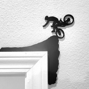 May include: Black metal wall decor featuring a silhouette of a person riding a mountain bike over a cliff edge.