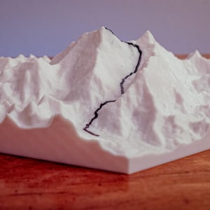 Puede incluir: Modelo blanco impreso en 3D de una cordillera con una línea oscura que representa un camino o río. El modelo está sobre una base cuadrada y descansa sobre una superficie de madera.