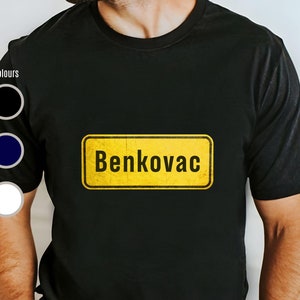 May include: Black t-shirt with a yellow sign that says "Benkovac" on it. The sign has a distressed look.