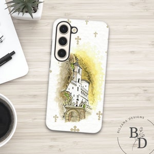May include: A white phone case with a watercolour illustration of a church or monastery on a cliff. The design has a yellow and brown backdrop, with small gold crosses on the white surface. The case has a camera module.