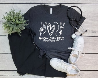 May include: A dark gray t-shirt with a white peace sign, heart, and medical symbol. The text on the shirt reads "Peace. Love. PICCS. Vascular Access Team."
