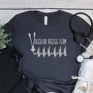 May include: A dark gray t-shirt with the text "Vascular Access Team" and a stylized heartbeat graphic. The shirt is made of a soft, comfortable material.