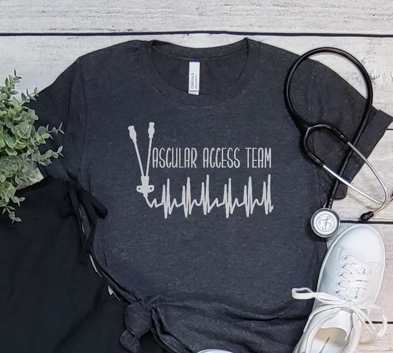 May include: A dark gray t-shirt with the text "Vascular Access Team" and a stylized heartbeat graphic. The shirt is made of a soft, comfortable material.