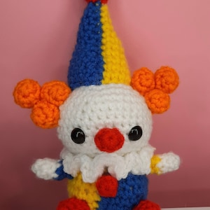 May include: A handmade crochet clown doll with a white head, a red nose, and black eyes. The clown has a blue, yellow, and red hat, orange hair puffs, and a blue, yellow, and red outfit. The doll has red pom-pom feet.