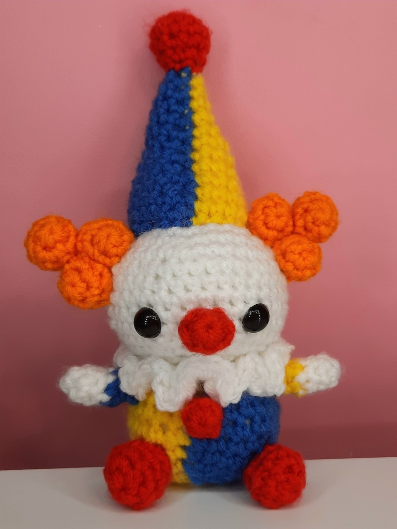 May include: A handmade crochet clown doll with a white head, a red nose, and black eyes. The clown has a blue, yellow, and red hat, orange hair puffs, and a blue, yellow, and red outfit. The doll has red pom-pom feet.