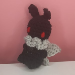 May include: A black crocheted plush toy with red eyes and a gray crocheted scarf. The toy is shaped like a rabbit with long ears.