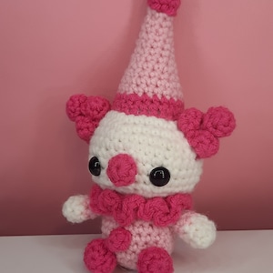 May include: A handmade crochet clown doll in shades of pink and white. The doll has a conical hat, ruffled collar, and large black eyes. The clown's features include pink pom-poms and a round nose. The doll is sitting on a white surface against a pink background.