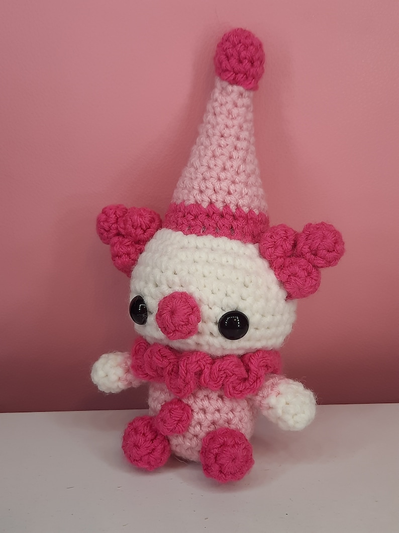May include: A handmade crochet clown doll in shades of pink and white. The doll has a conical hat, ruffled collar, and large black eyes. The clown's features include pink pom-poms and a round nose. The doll is sitting on a white surface against a pink background.