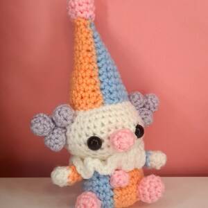 May include: A handmade crochet clown doll with a colorful design. The clown features a white body, a ruffled collar, and a tall hat in orange, blue, and pink. It has black button eyes, a pink nose, and lavender hair puffs. The clown is sitting on a white surface.