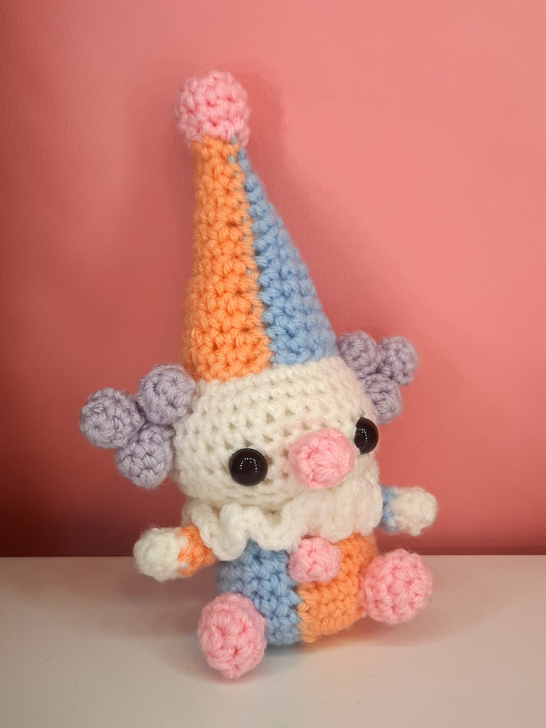 May include: A handmade crochet clown doll with a colorful design. The clown features a white body, a ruffled collar, and a tall hat in orange, blue, and pink. It has black button eyes, a pink nose, and lavender hair puffs. The clown is sitting on a white surface.