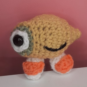 Pode incluir: Um brinquedo de pelúcia de crochê em forma de olho de desenho animado com corpo amarelo, pálpebras verdes, esclera branca e pupila preta. O brinquedo tem dois pés de crochê laranja e branco.