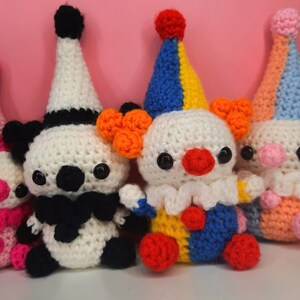 May include: A group of four handmade crochet clown dolls. Each doll has a white head, a colorful hat, and a round red nose. The clowns have black eyes and are wearing different colored outfits, including pink, black, blue, and orange.