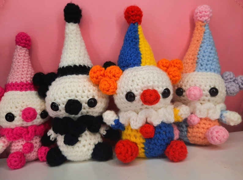 May include: A group of four handmade crochet clown dolls. Each doll has a white head, a colorful hat, and a round red nose. The clowns have black eyes and are wearing different colored outfits, including pink, black, blue, and orange.