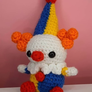 May include: A handmade crochet clown doll with a white head, a red nose, and black eyes. The clown has a blue, yellow, and red hat, orange hair, and a blue and yellow outfit. The doll is sitting on a white surface against a pink background.