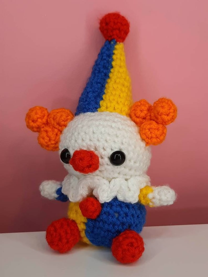 May include: A handmade crochet clown doll with a white head, a red nose, and black eyes. The clown has a blue, yellow, and red hat, orange hair, and a blue and yellow outfit. The doll is sitting on a white surface against a pink background.