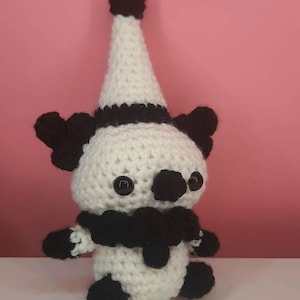 May include: A handmade crochet clown doll in white and black. The doll has a white cone hat with a black band and a black pom-pom on top. It has black button eyes, black ears, and a black ruffled collar. The doll's arms and feet are also black.